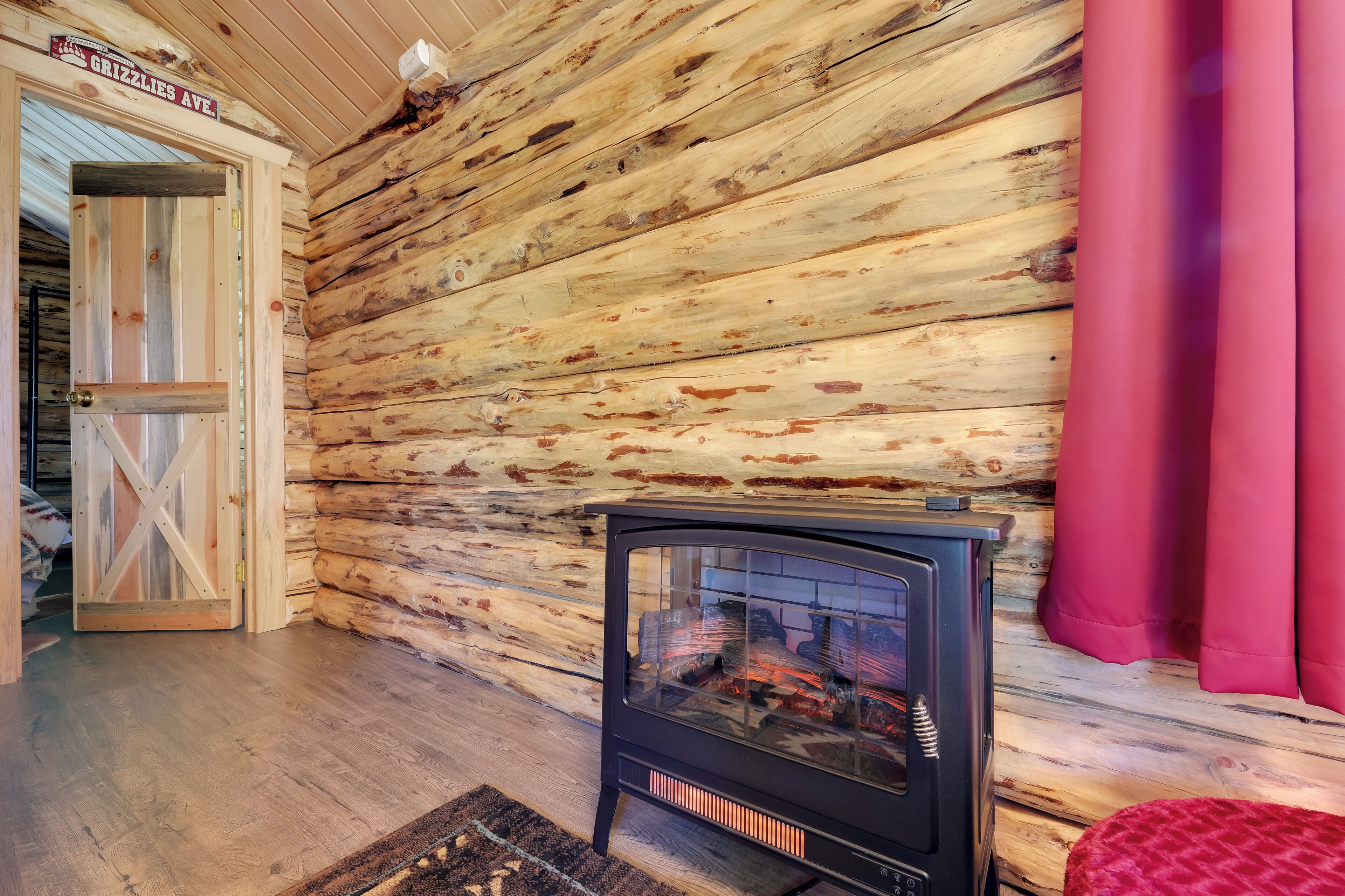 A rustic cabin interior with a wooden log wall, a black wood stove, and a red curtain.