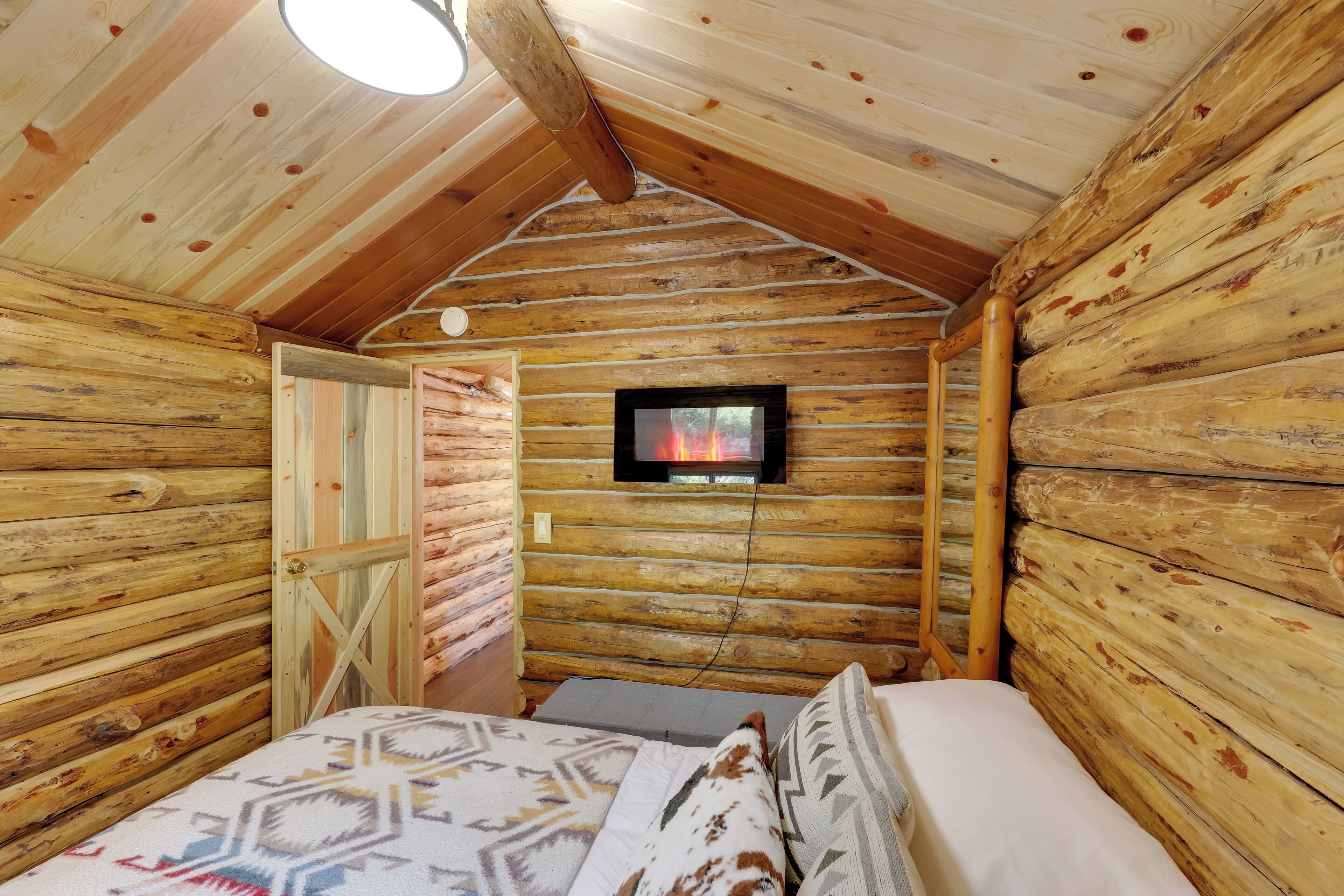 A log cabin room with a bed, a television mounted on the wall, and a wooden ceiling.