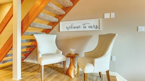 A loft-style room with a wooden staircase, two white armchairs, a small wooden table, and a wall sign that reads "welcome to ours". - Waterfront Community: Lake Winnipesaukee Retreat! (Gilford)