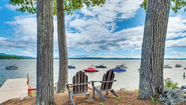 A lakeside view with two Adirondack chairs facing a dock and boats on the water. - Waterfront Community: Lake Winnipesaukee Retreat! (Gilford)