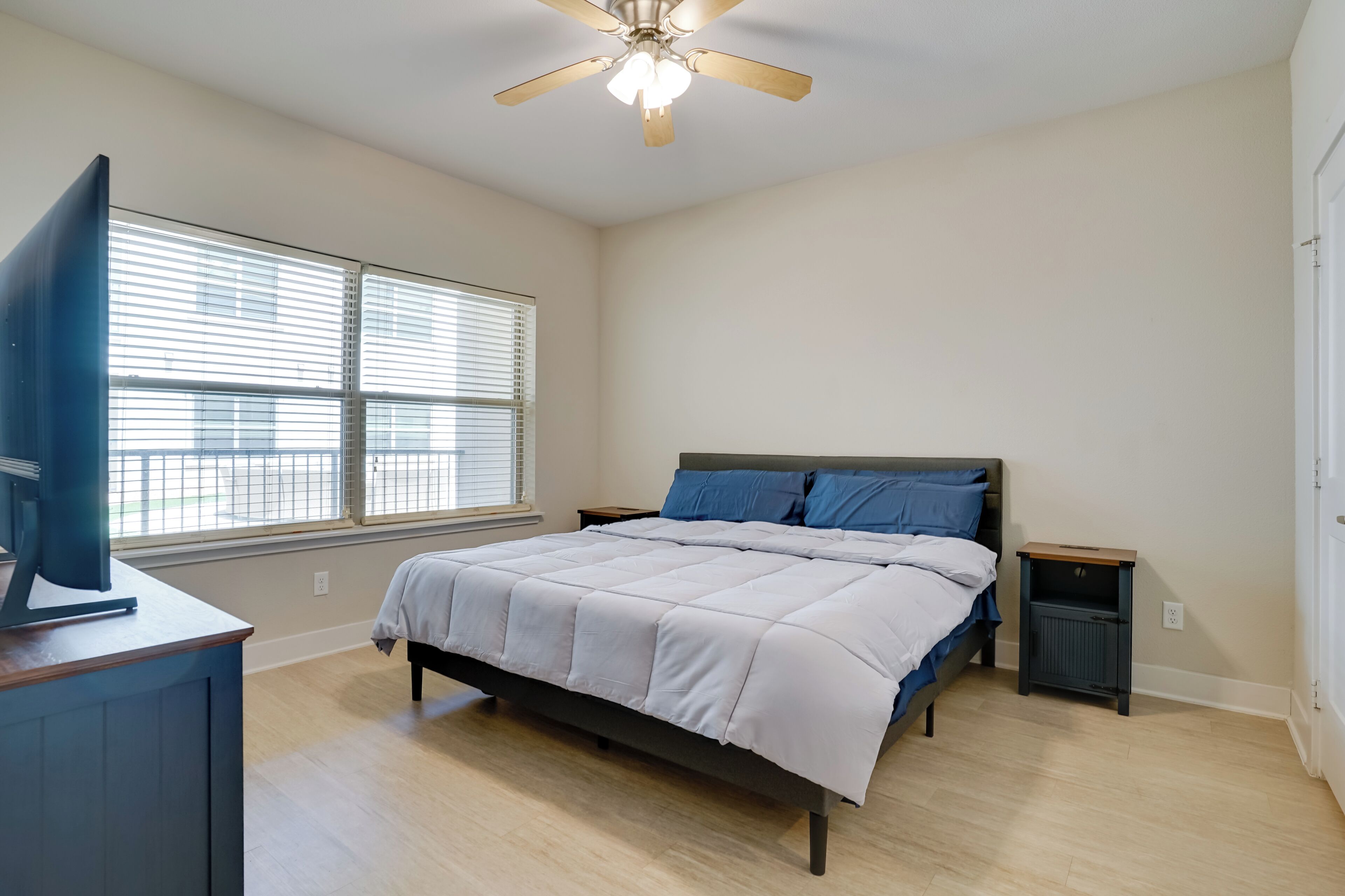 Un dormitorio con cama, televisor sobre un mueble bajo, ventilador de techo y ventana con persianas.