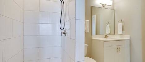 A bathroom with white tiles, a shower with a handheld showerhead, a toilet, and a vanity with a mirror and towel rack.