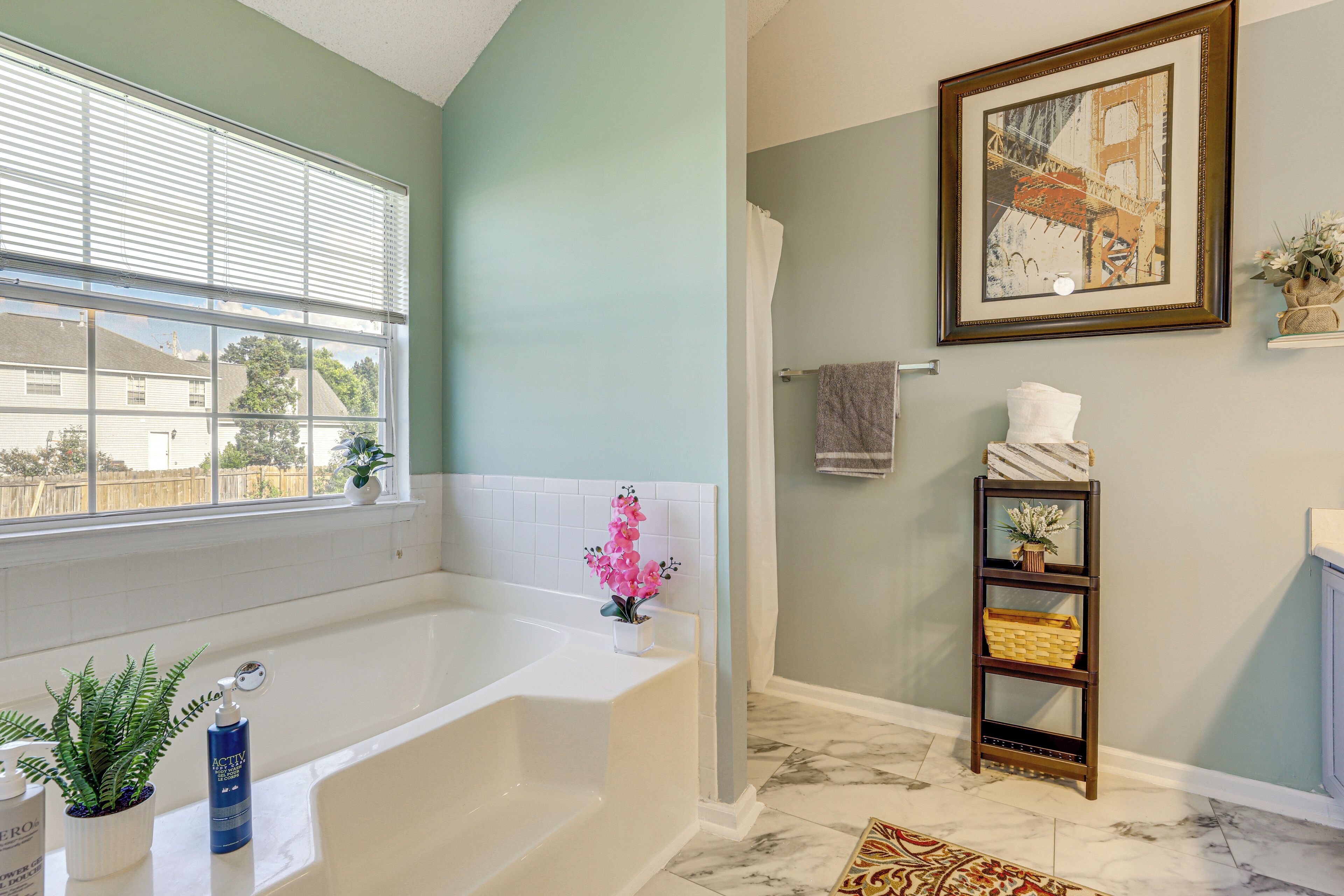 A bathroom with a bathtub, a window with blinds, a shower curtain, a towel rack, a shelf with decorative items, and a framed picture on the wall.