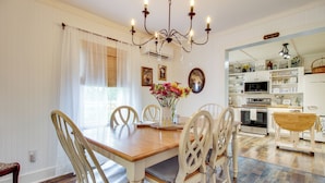 A dining area with a wooden table, white chairs, a chandelier, and a kitchen in the background. - Shepard Cottage Near Wilmington & Wedding Venue (Burgaw)
