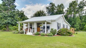 A white house with a porch, surrounded by a well-maintained lawn and trees. - Shepard Cottage Near Wilmington & Wedding Venue (Burgaw)
