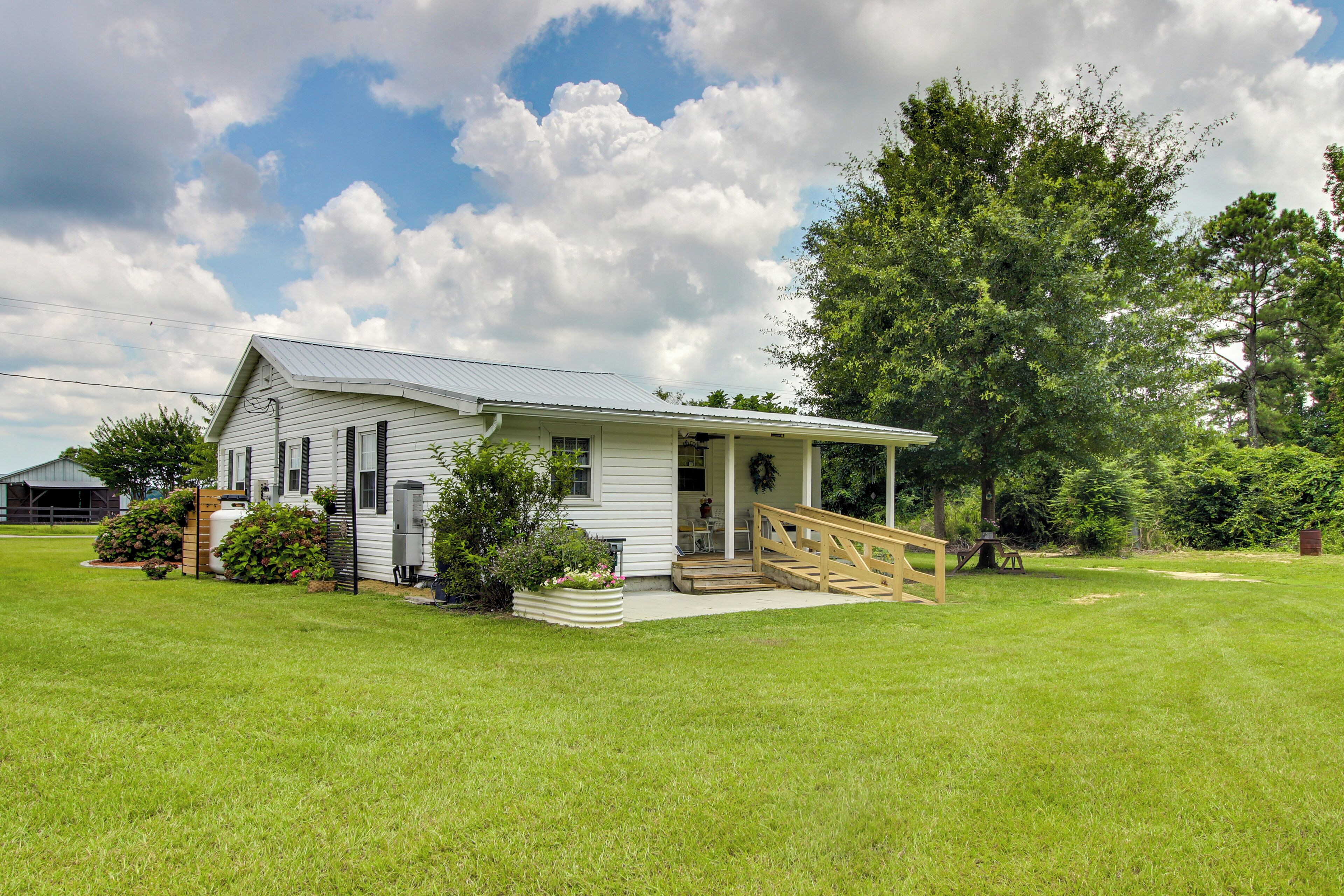 A white single-story house with a porch, a small garden, and a large tree in the background.