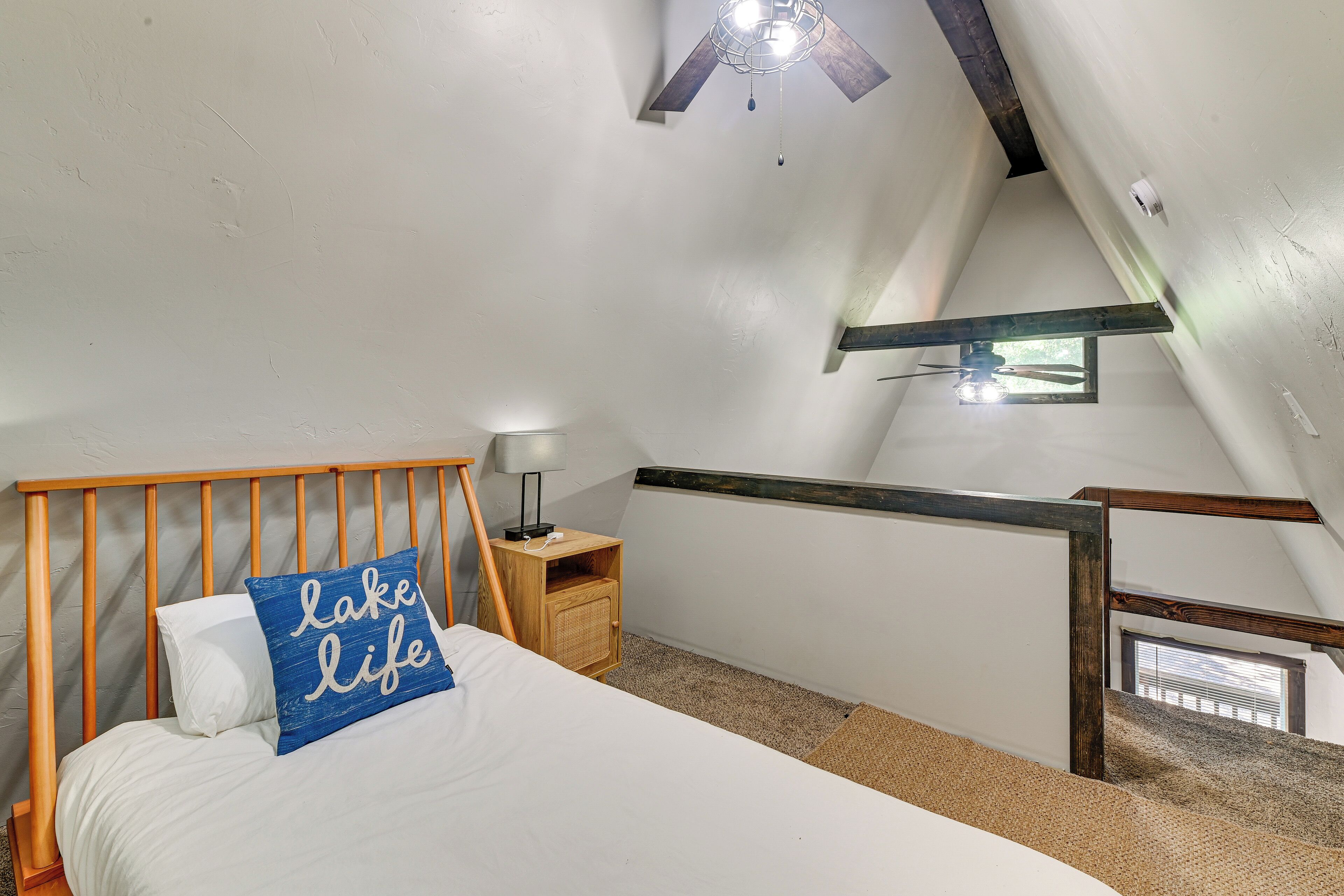 A bedroom with a bed, a nightstand, a lamp, a ceiling fan, a window, and a wooden staircase.