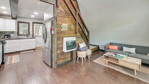 A loft-style living space with a kitchen, wooden beams, a fireplace, and a sofa with cushions.
