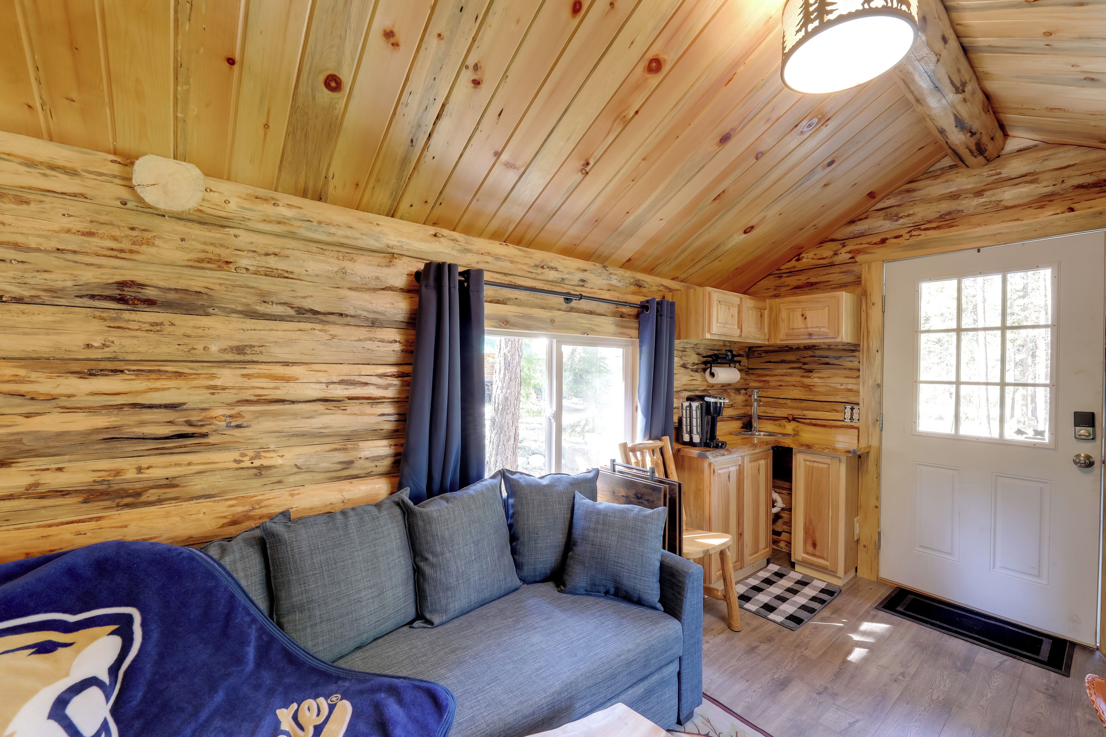 A cozy cabin interior with a wooden wall, a sofa with cushions, a coffee station, and a door leading outside.