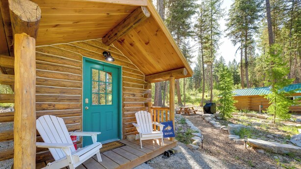 A log cabin with a green door, two white Adirondack chairs, and a stone bench outside.