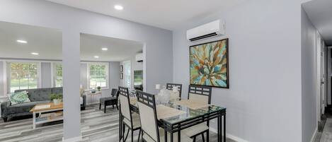 A modern dining area with a glass-top table and black chairs, a grey sofa, and a colorful abstract painting on the wall.