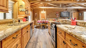 A log cabin interior with wooden walls, a kitchen with granite countertops, a dining area with a red tablecloth, and a large TV on the wall. - Near Fishing & Hiking: Pinetop Log Cabin w/ Deck! (Pinetop)