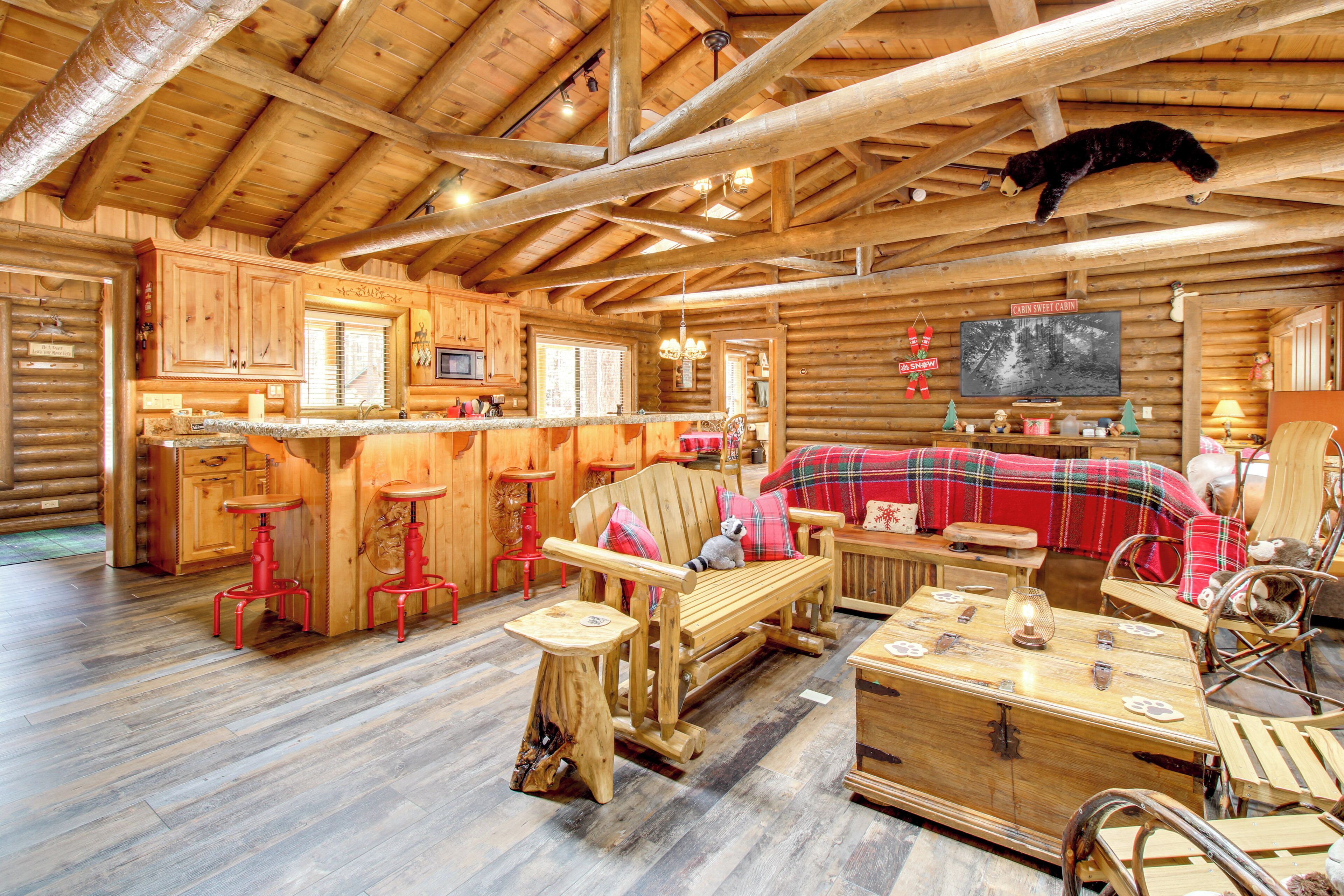 A log cabin interior with a wooden dining table, a bench with a blanket, a television, and a bear decoration on the ceiling.