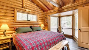 A log cabin bedroom with a plaid bedspread, two bedside lamps, a bench, and a window with blinds. - Near Fishing & Hiking: Pinetop Log Cabin w/ Deck! (Pinetop)
