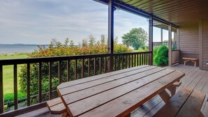 A wooden deck with a picnic table and benches, overlooking a body of water and greenery. - Community Boat Dock: Lake Memphremagog Condo! (Newport Center)