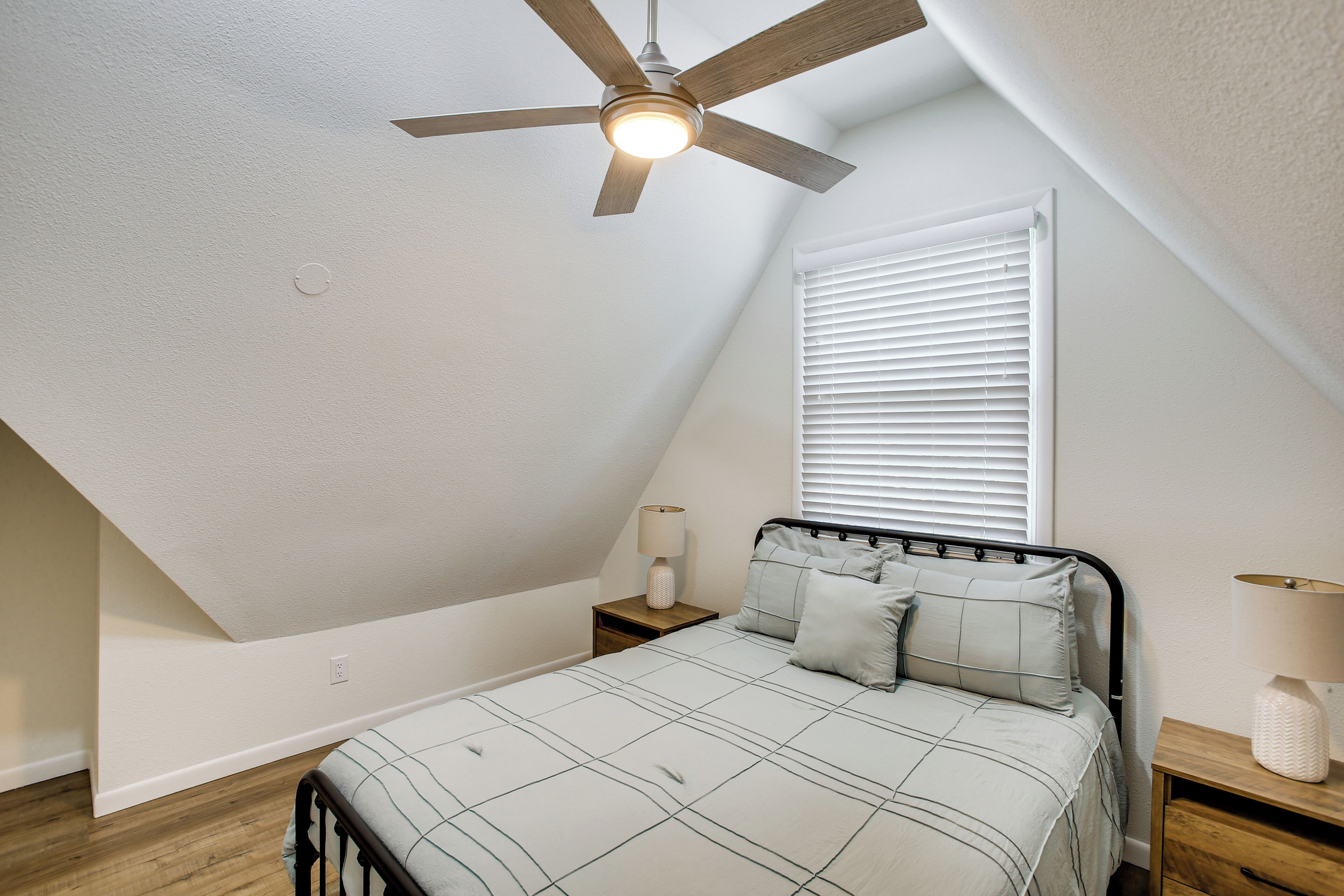 A bedroom with a sloped ceiling, a ceiling fan, a bed with a headboard, a nightstand, a lamp, and a window with blinds.
