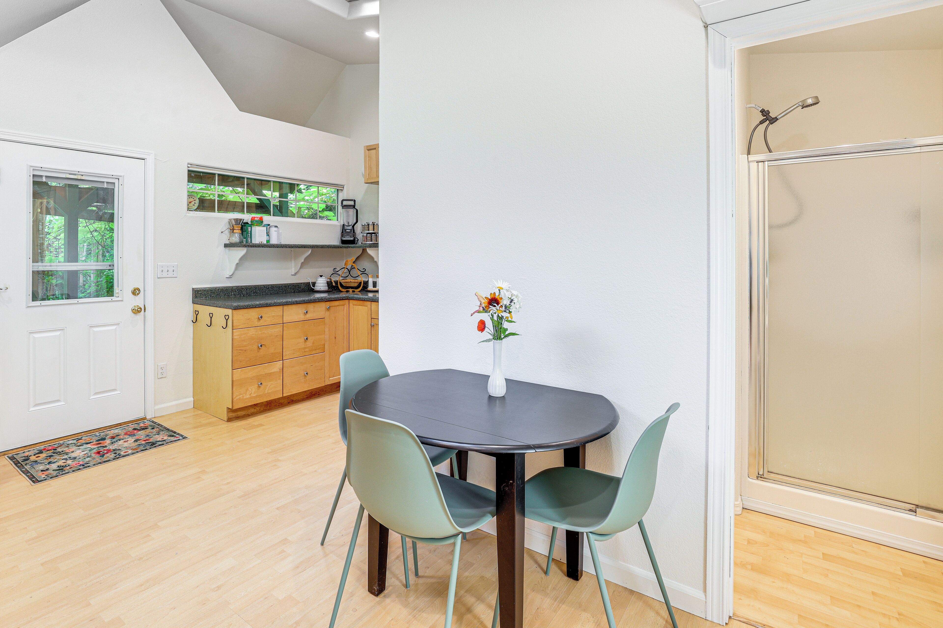 A dining area with a round table, four chairs, and a vase with flowers. There is a kitchen in the background with wooden cabinets and a window.