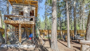 Una cabaña de madera rústica con balcón, rodeada de árboles altos y terreno de tierra.