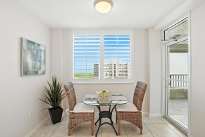 A dining area with a glass table and wicker chairs, a potted plant, and a window with blinds.