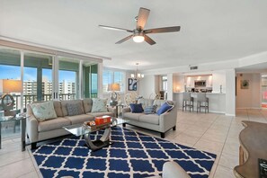 A modern living room with a sofa, coffee table, and patterned rug. A ceiling fan is visible, and the room has large windows.
