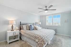 A bedroom with a bed, nightstand, lamp, ceiling fan, window with blinds, and a view of buildings outside.