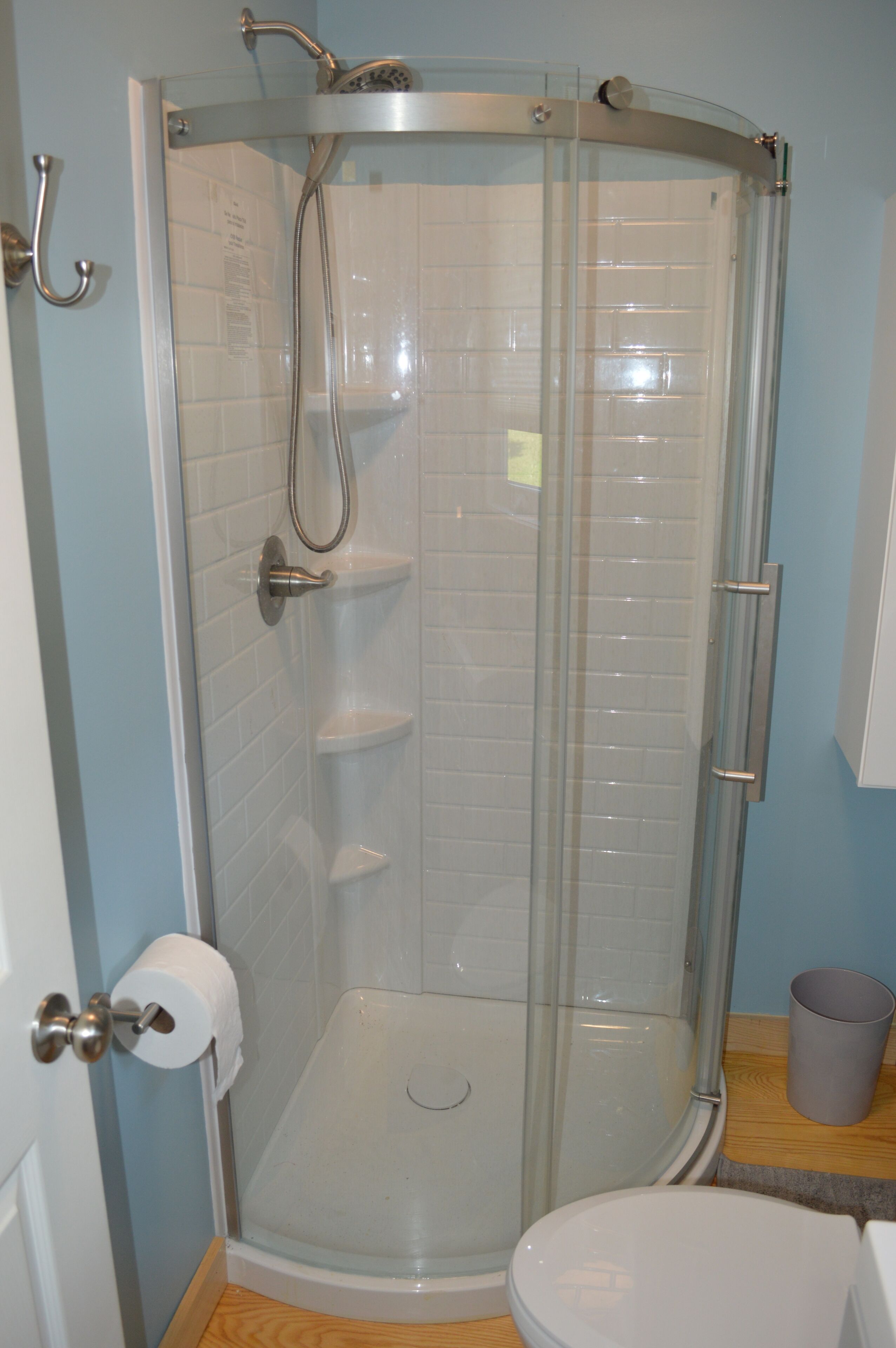A glass-enclosed shower stall with a toilet and a trash can in a bathroom.