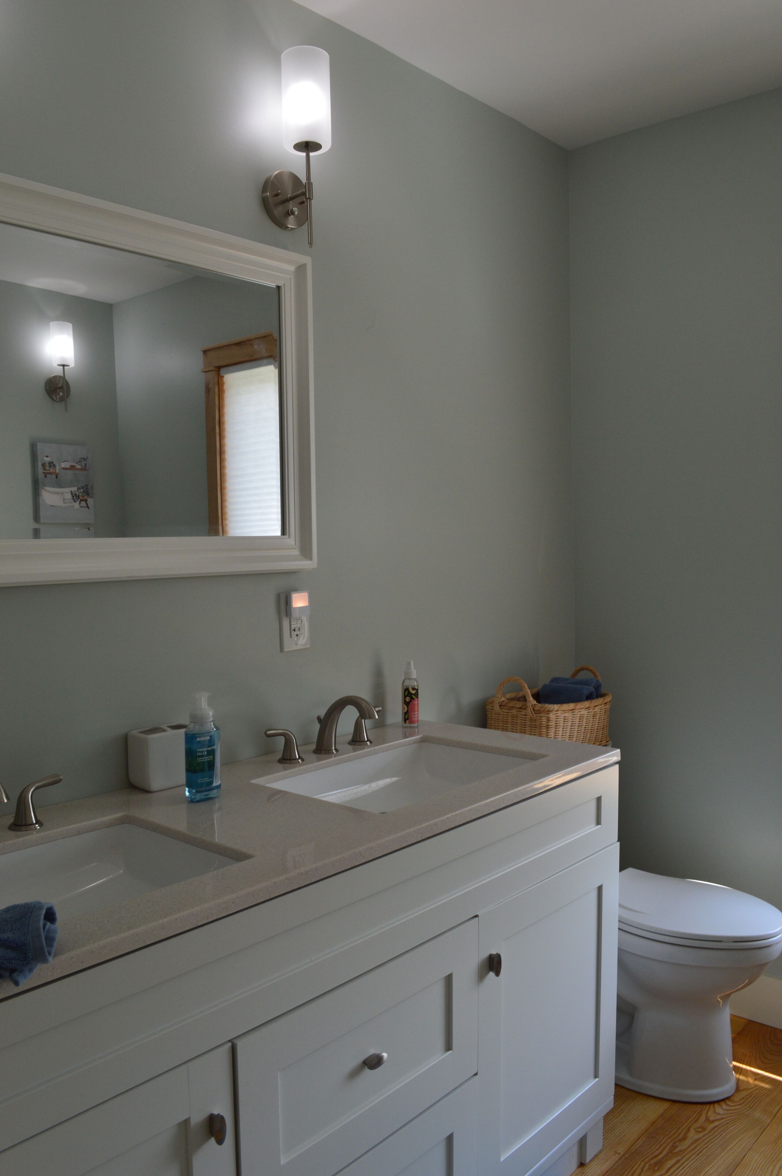 A bathroom with two sinks, a mirror, a toilet, and a basket on the floor.