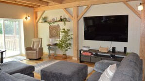 A living room with a large flat-screen TV, grey sofas, a wooden ceiling, and a small table with a lamp and plant.