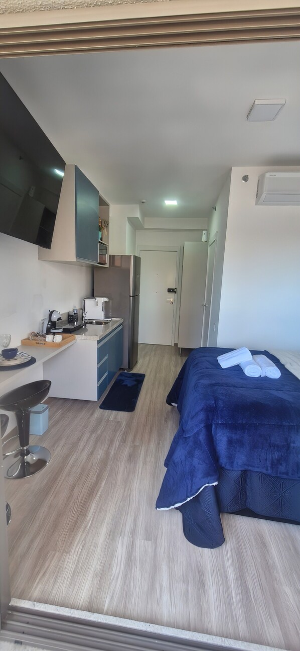 A compact hotel room with a kitchenette, a bed with a dark blue blanket, a small seating area, and a door leading to another room.