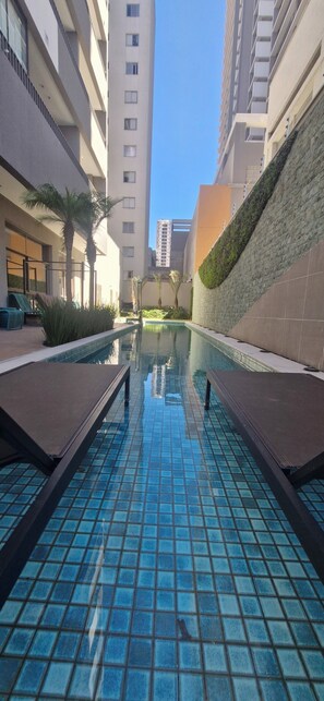 A swimming pool with tiled flooring, surrounded by lounge chairs and palm trees, with high-rise buildings in the background.