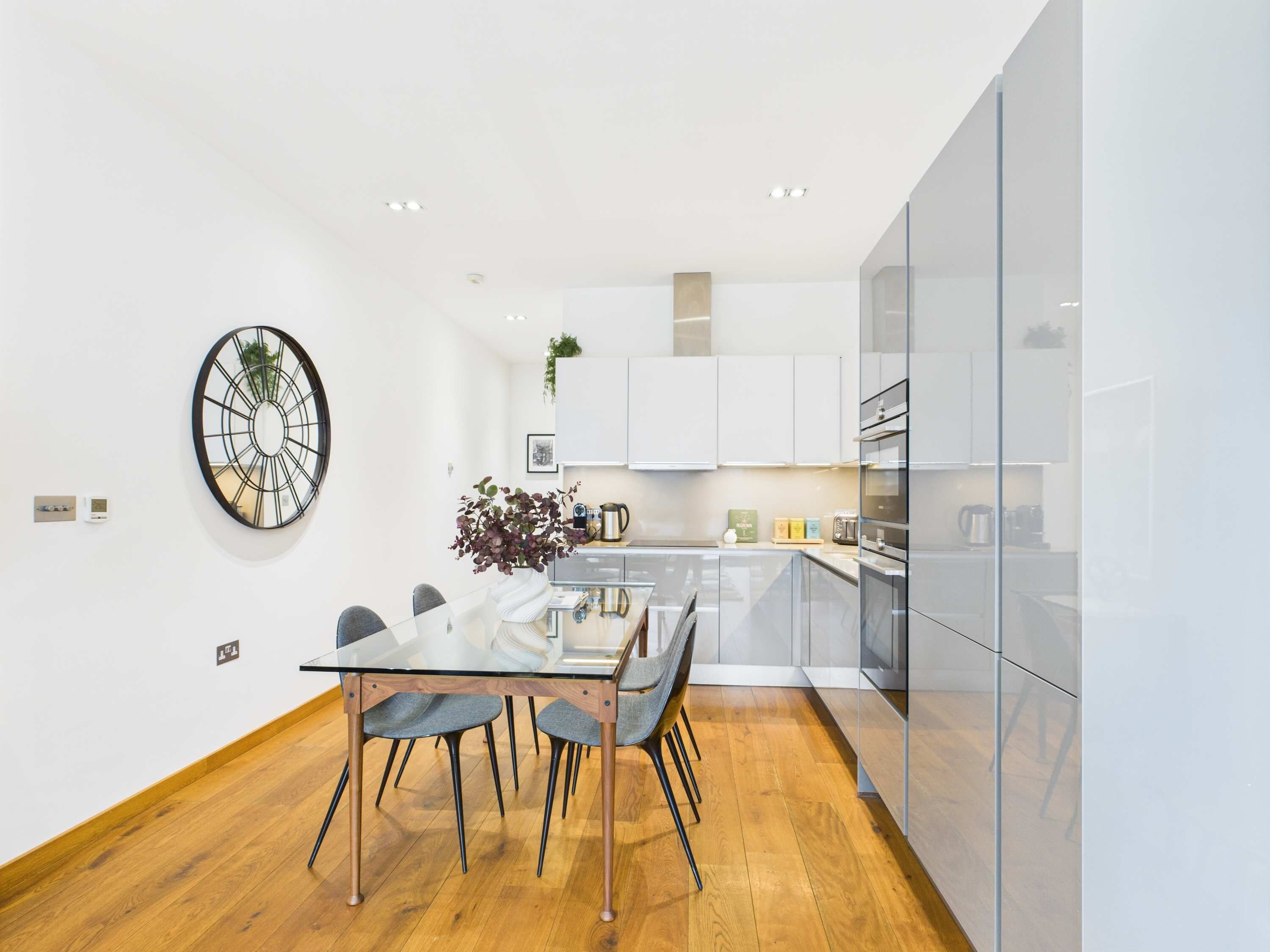 Una cocina moderna con armarios blancos, una mesa de comedor de vidrio y suelo de madera.