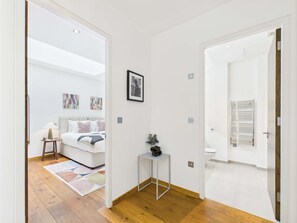 Ein modernes Hotelzimmer mit Holzboden, einem Bett mit weißem Kopfteil, einem kleinen Nachttisch mit Lampe und einem gerahmten Bild an der Wand.