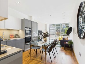 A modern kitchen with a glass dining table, grey cabinets, and a large window offering a view of the outdoors.