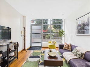 A modern living room with a sofa, coffee table, and TV. There is a large window with a view of the outside. - Modern Apartment in Camden Warehouse Conversion (Greater London)