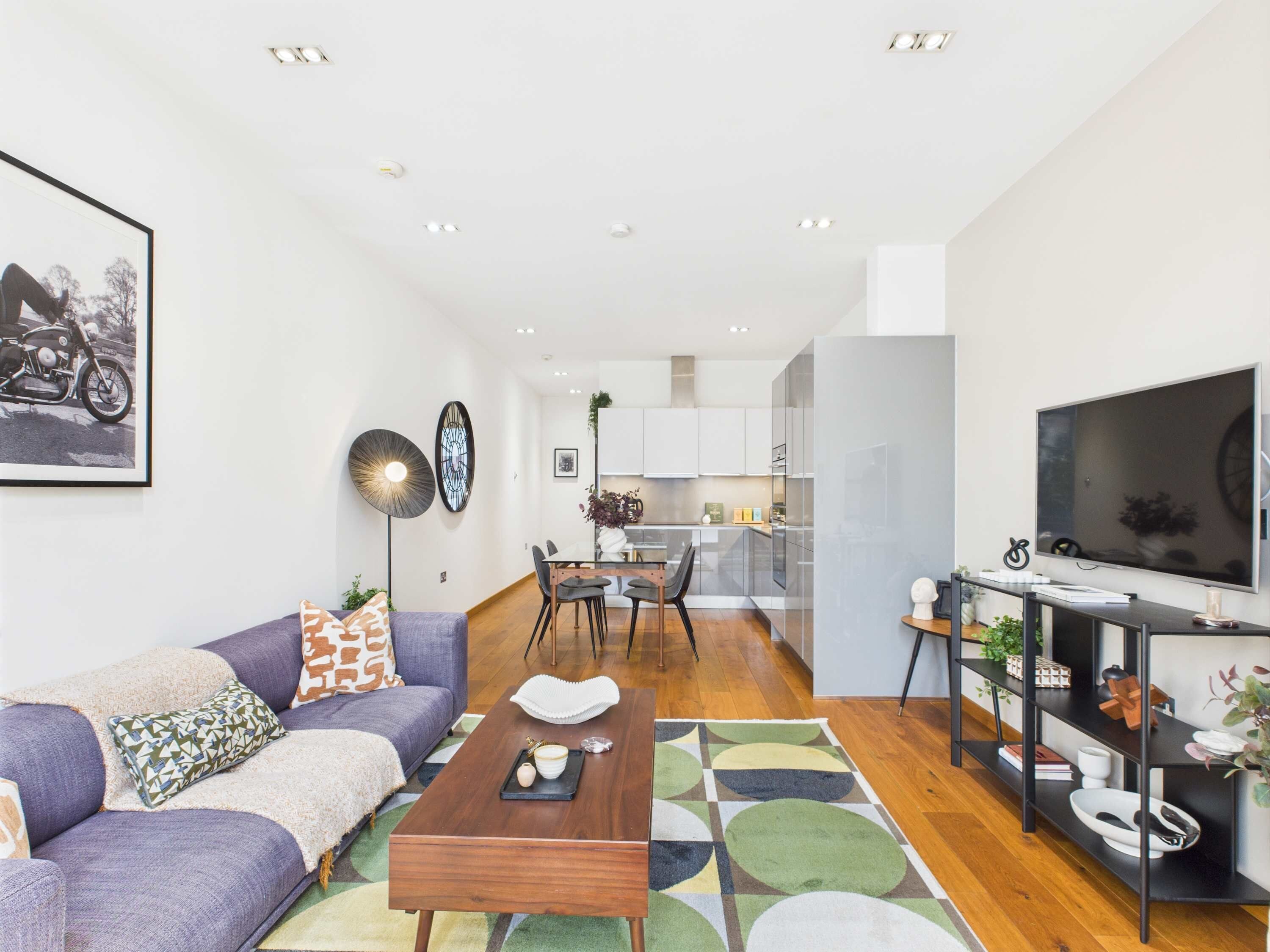 A modern living room with a sofa, coffee table, and a TV. There is a dining area with a table and chairs in the background.