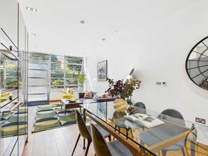 A modern living room with a glass table, comfortable seating, and a large window offering a view of greenery.