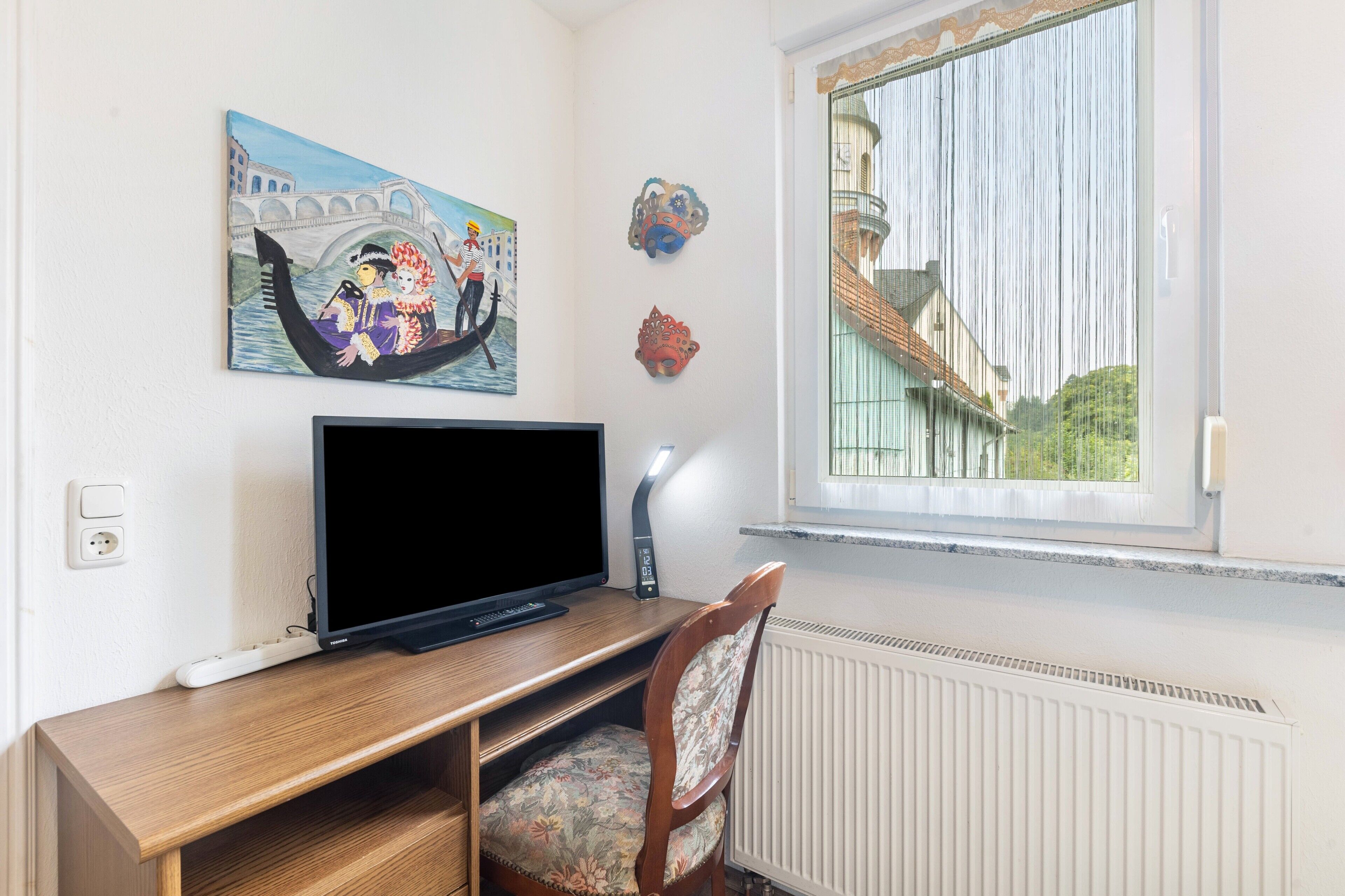 A room with a TV on a wooden stand, a chair, a painting of a gondola, and a window with a view of a building.
