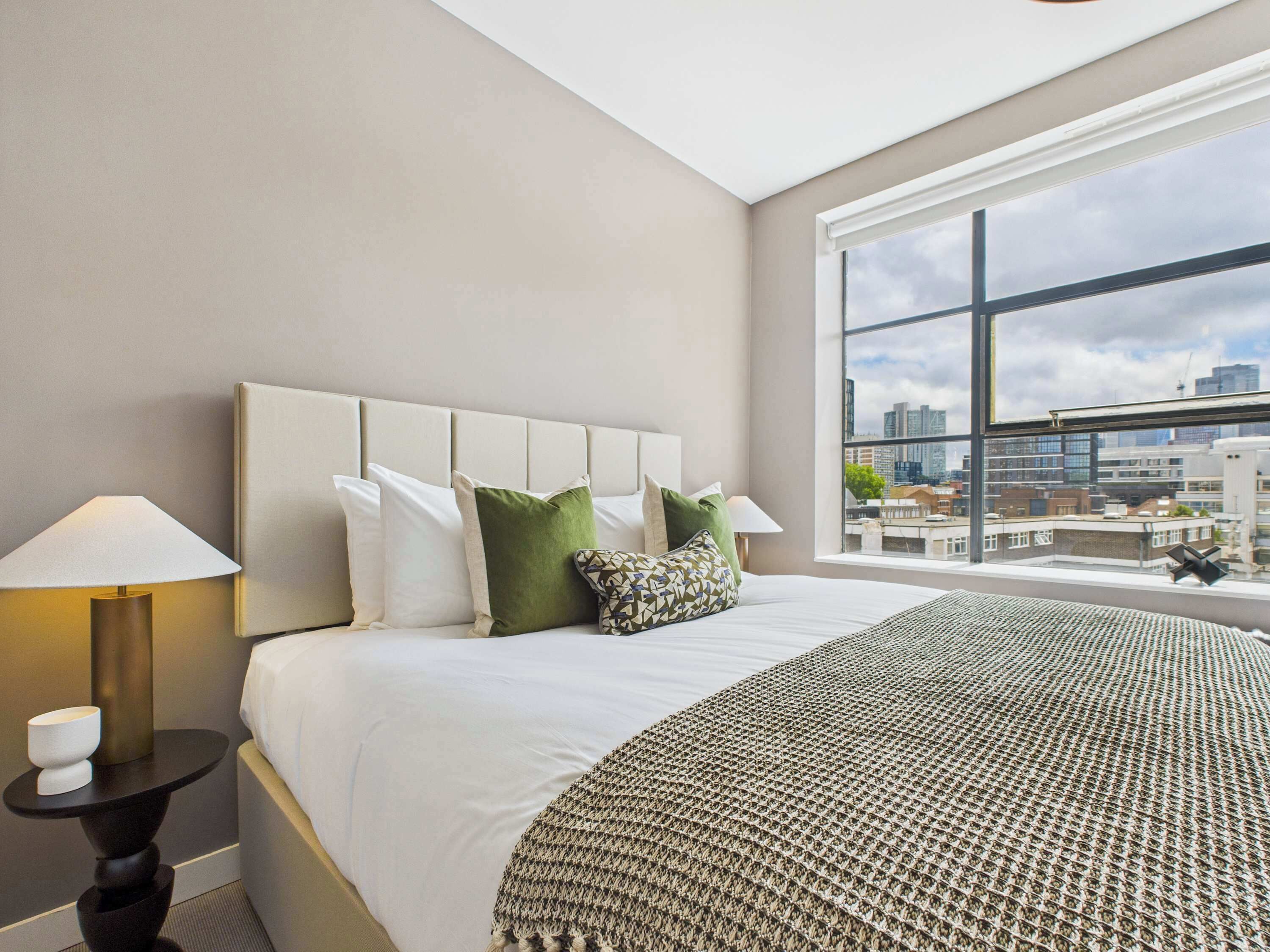 Ett modernt hotellrum med en stor säng, ett nattduksbord, en lampa och utsikt över staden genom ett stort fönster.