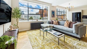 A modern living room with a grey sofa, a glass coffee table, and a large window offering a city view.