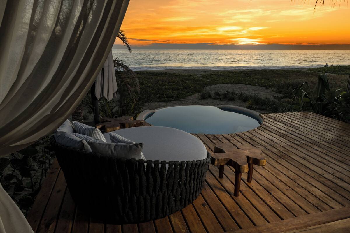 Una terraza de madera con una zona de estar circular y un jacuzzi, con vistas a una playa al atardecer.