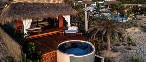 A thatched-roof hut with a wooden deck, a hot tub, and a pool area with lounge chairs.
