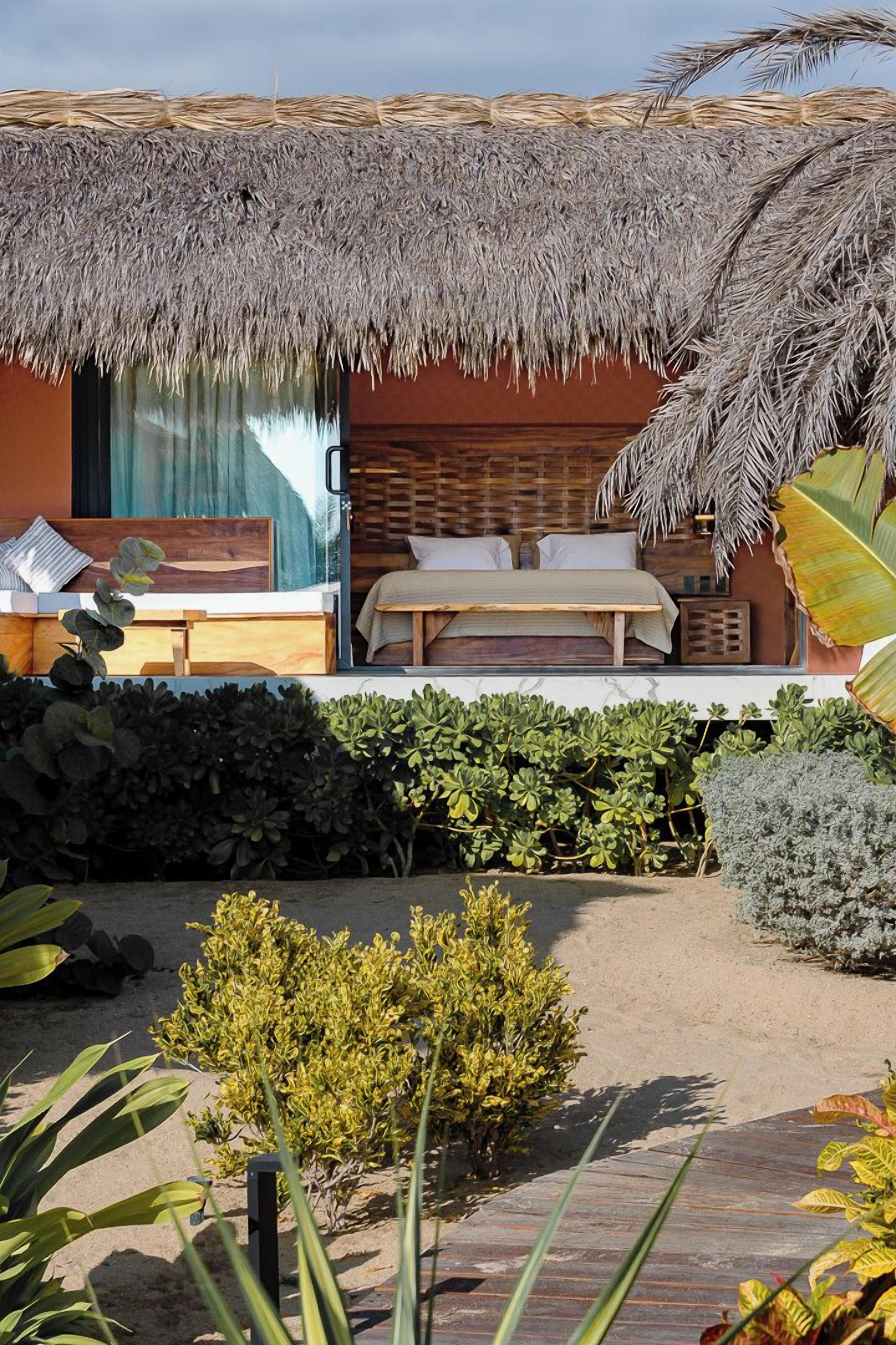 Una cabaña con techo de paja que incluye una cama, un pequeño espacio de descanso y un jardín con diversas plantas.