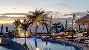 Ein geschwungener Infinity-Pool mit Liegestühlen und Sonnenschirmen, umgeben von Palmen und Blick auf einen Sonnenuntergang.