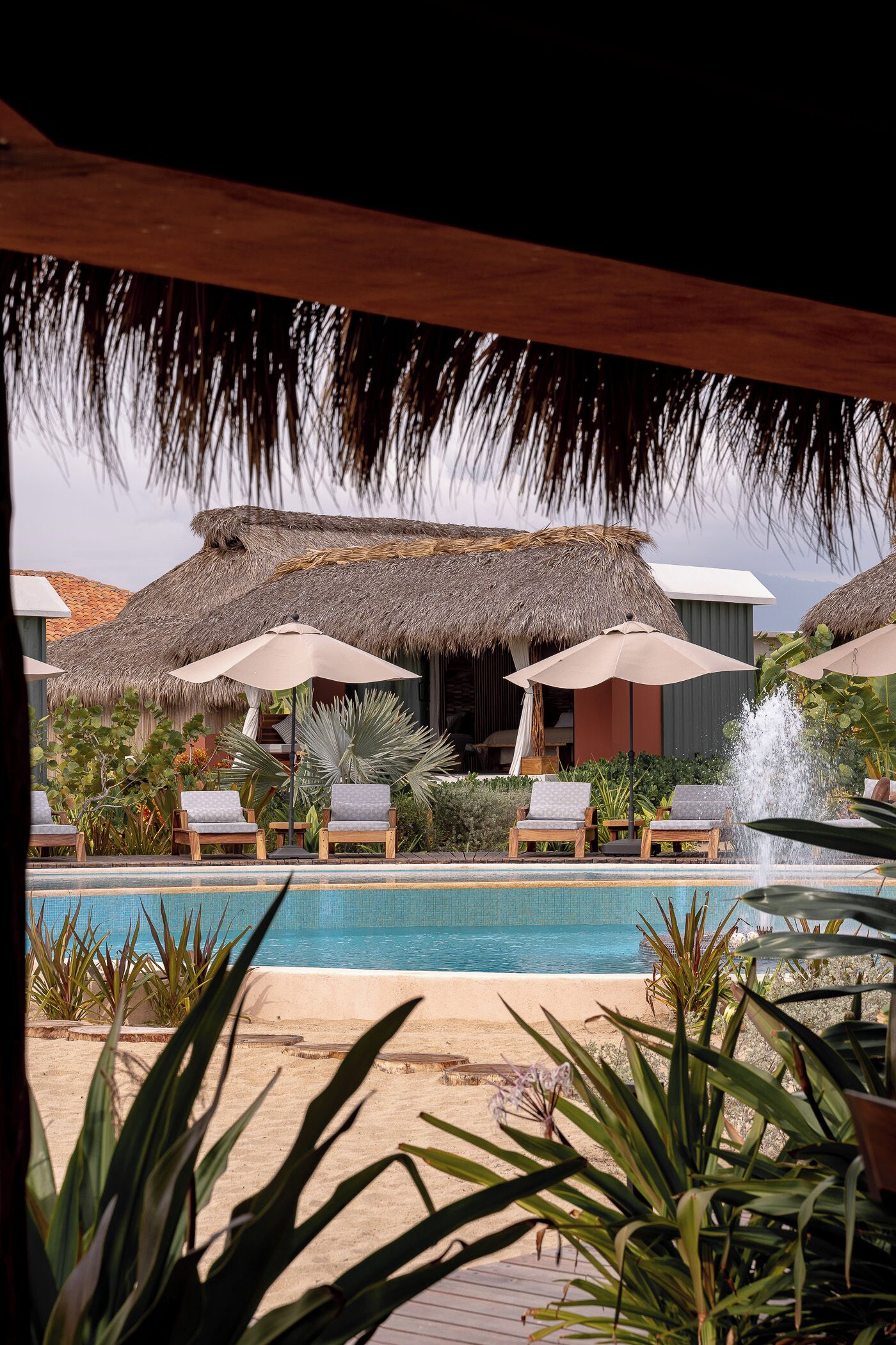 A pool area with lounge chairs and umbrellas, a thatched-roof building, and a water feature.