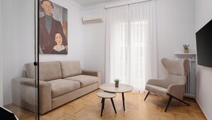 A living room with a beige sofa, a round coffee table, and a beige armchair. There is a painting on the wall, a window with white curtains, and an air conditioning unit.