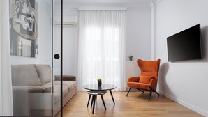 A modern living room with a grey sofa, an orange armchair, a small round table with a plant, and a flat-screen TV mounted on the wall.