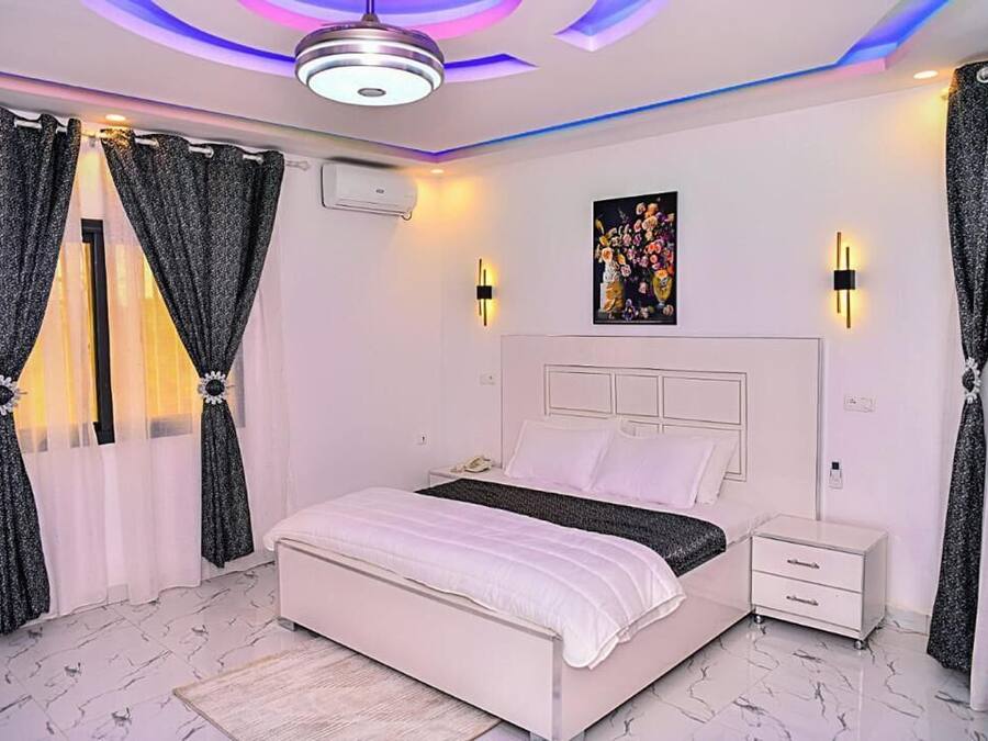 Un dormitorio con cama blanca, mesitas de noche, lámpara de techo y cortinas decorativas.