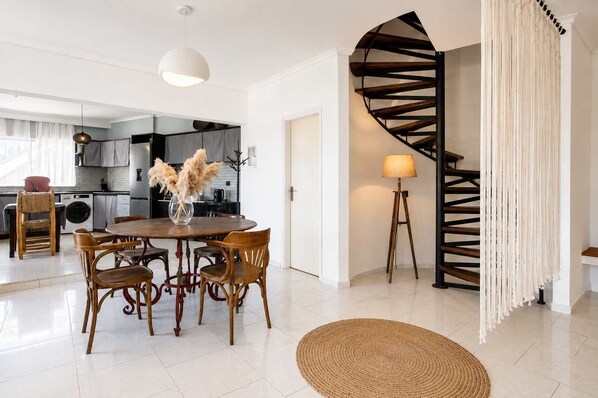 A modern kitchen with white cabinets, a dining area with a round wooden table and chairs, a spiral staircase, and a large window.