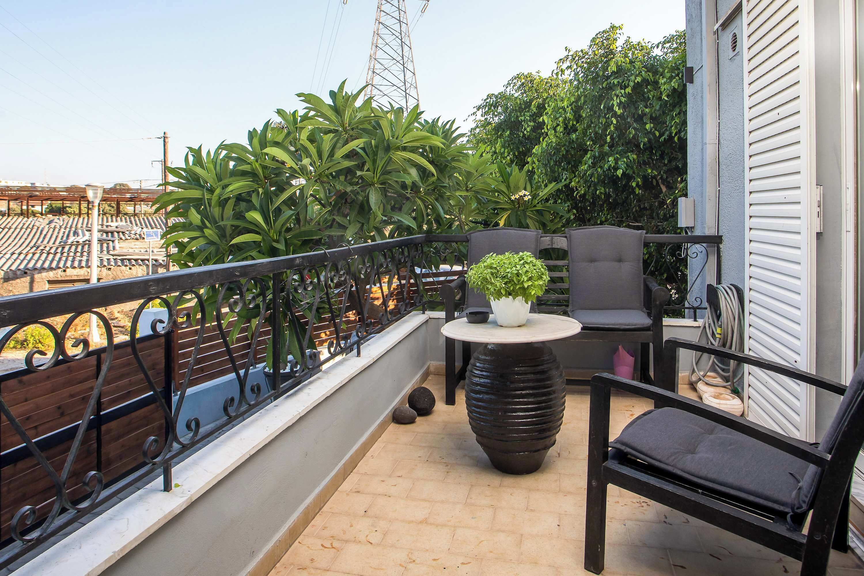 Un balcon doté d’une balustrade en métal noir, une table ronde avec un pot de fleurs, une chaise noire et une vue sur un paysage urbain verdoyant.