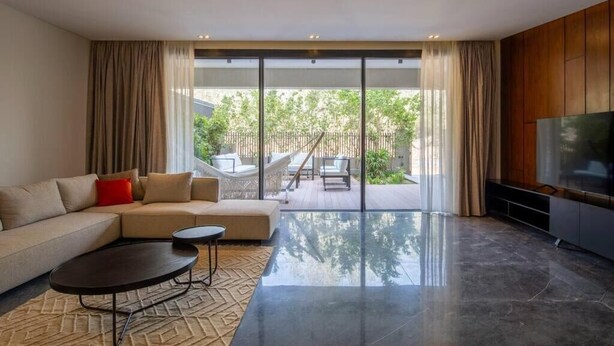 A modern living room with a sofa, a coffee table, and a TV. There is a sliding glass door leading to an outdoor area with a hammock and a wooden deck. - Kennah Catalina | West Golf Extension, New Cairo (New Cairo)